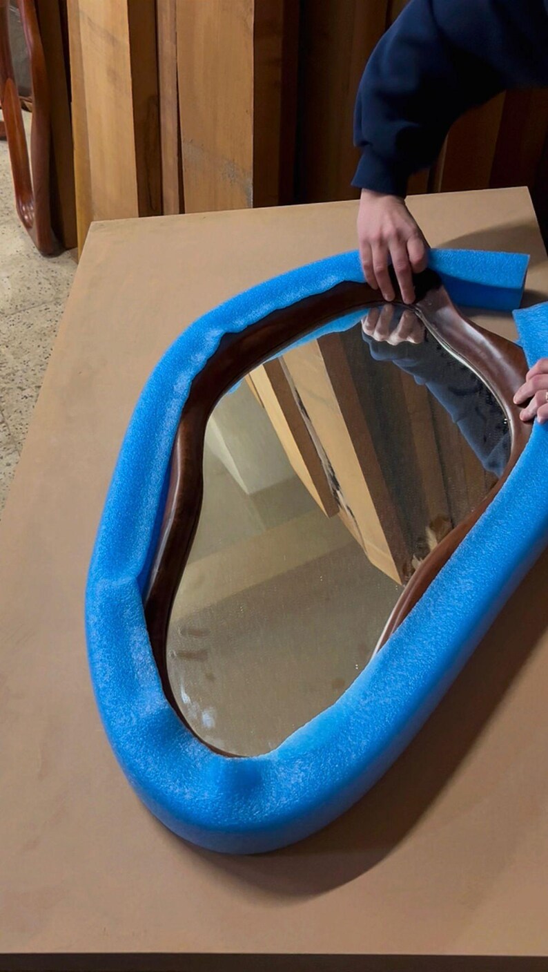May include: An irregular, organic-shaped mirror with a dark brown wooden frame. The mirror is partially wrapped in bright blue foam protective padding, which contours around the curved edges of the wooden frame to provide cushioning.