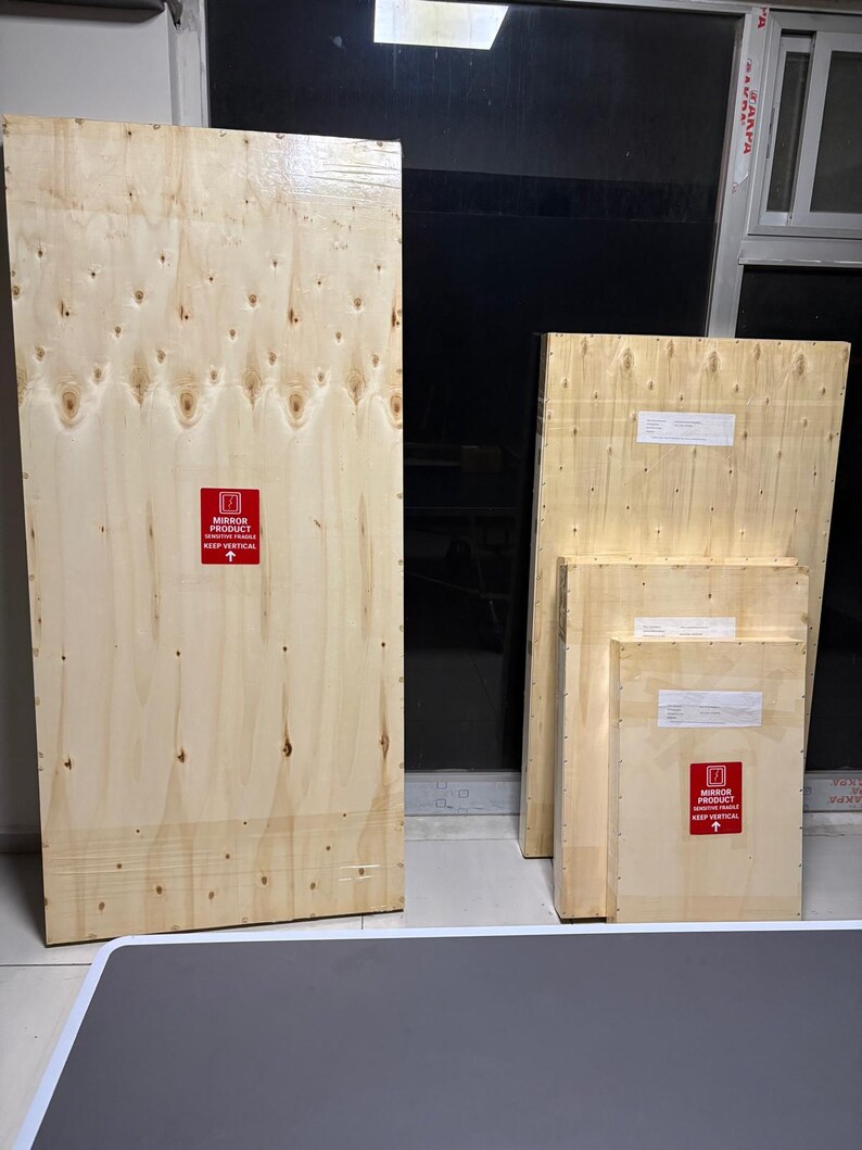 May include: Four rectangular wooden shipping crates of varying sizes, each constructed from light-coloured plywood. Each crate features a red square label with white text reading MIRROR PRODUCT SENSITIVE FRAGILE KEEP VERTICAL and an upward arrow.