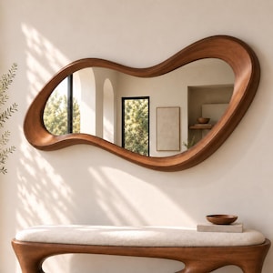 May include: A large, wall-mounted mirror featuring an irregular, organic, wavy wooden frame with a smooth, warm brown finish. The reflective surface shows a room interior with arched architectural elements, a framed picture, and a small bowl.