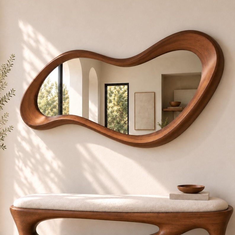 May include: A large, wall-mounted mirror featuring an irregular, organic, wavy wooden frame with a smooth, warm brown finish. The reflective surface shows a room interior with arched architectural elements, a framed picture, and a small bowl.