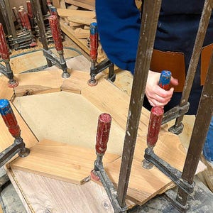 May include: A wooden frame assembly held together by multiple metal bar clamps with red and blue handles. The frame consists of several light brown wooden segments arranged in a geometric shape, secured onto a flat base during the construction process.