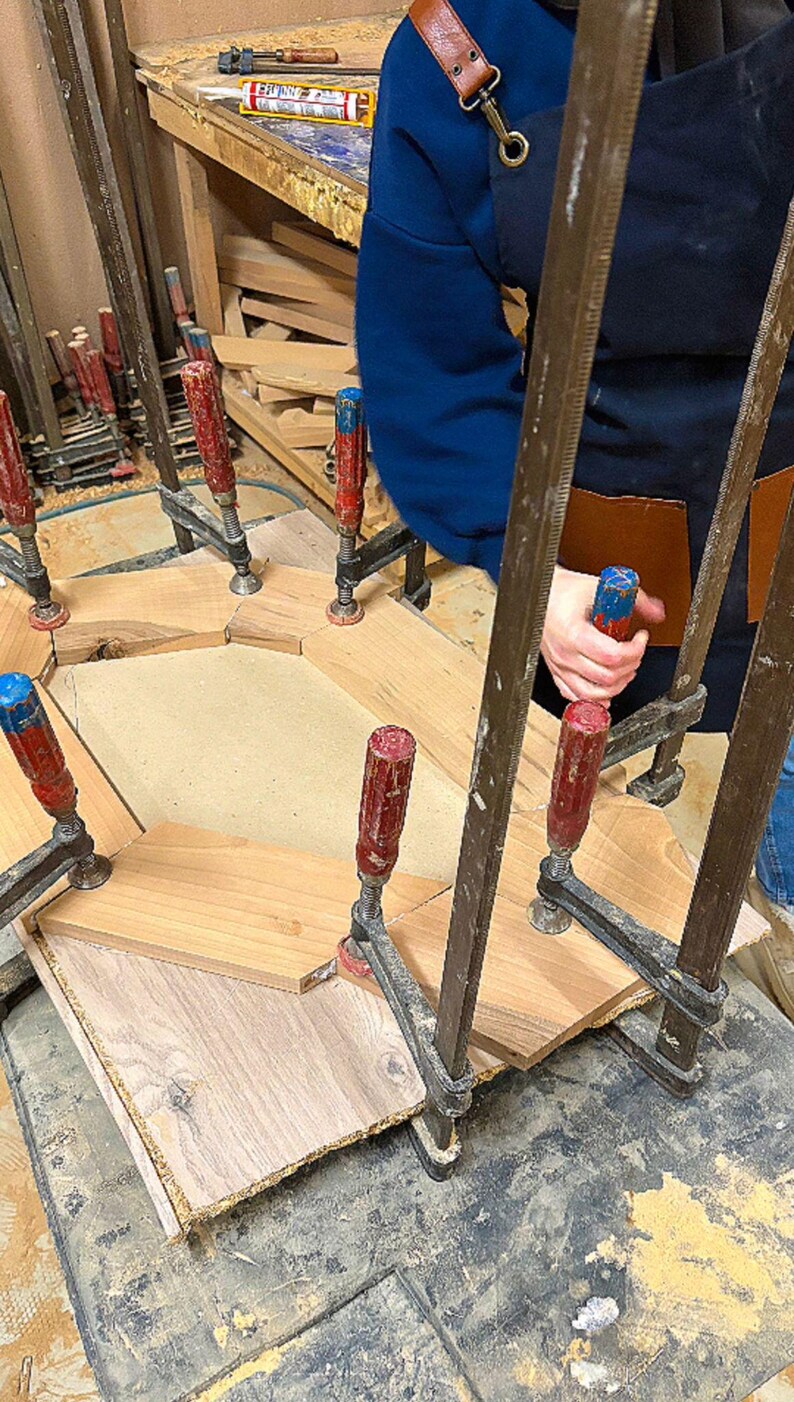 May include: A wooden frame assembly held together by multiple metal bar clamps with red and blue handles. The frame consists of several light brown wooden segments arranged in a geometric shape, secured onto a flat base during the construction process.