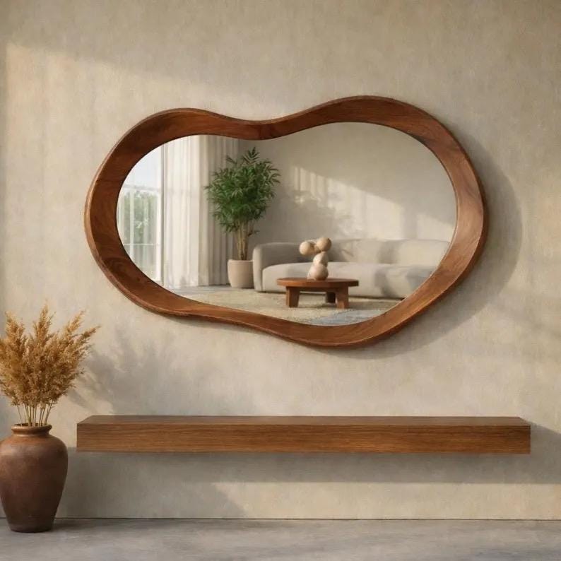 May include: A wall-mounted mirror with an irregular, organic-shaped wooden frame. The mirror reflects a room containing a sofa, a small wooden coffee table, and a potted plant. Below the mirror is a long, rectangular wooden floating shelf.