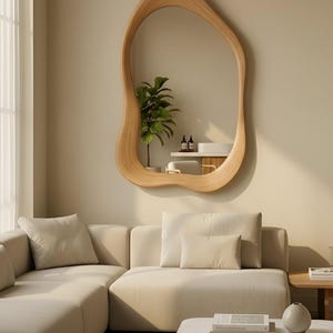 Puede incluir: Un espejo de pared con un marco de madera irregular, org&aacute;nico y ondulado con un acabado de veta de madera natural clara. El espejo refleja una planta verde en maceta, un estante blanco y dos botellas peque&ntilde;as, mostrando una silueta curva.