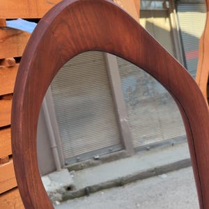 Puede incluir: Un espejo de pared con un marco de madera de color marr&oacute;n oscuro liso, que presenta una forma org&aacute;nica, asim&eacute;trica y curva. La superficie de vidrio reflectante est&aacute; encajada en el borde de madera maciza, mostrando una veta natural.