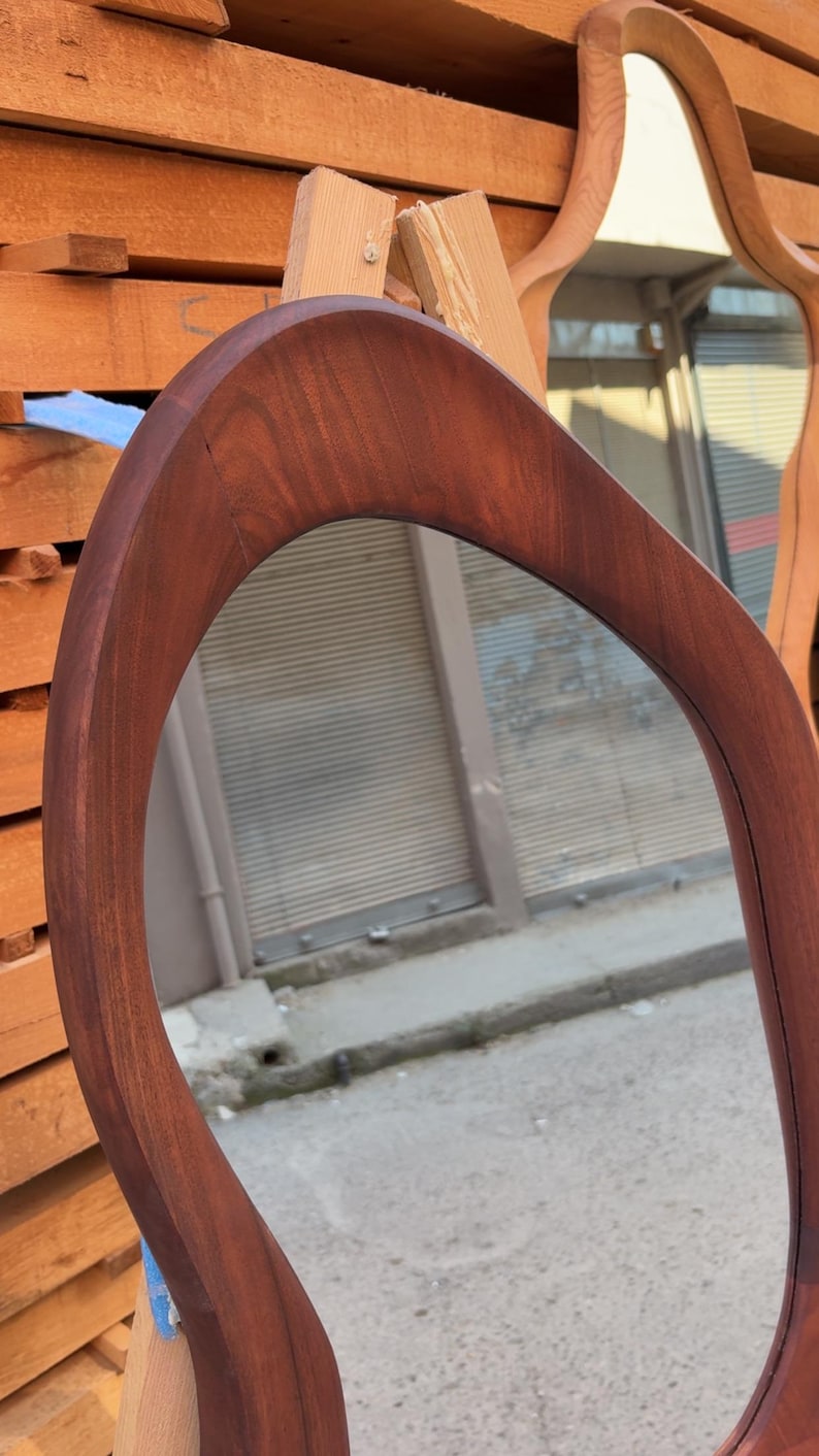 Puede incluir: Un espejo de pared con un marco de madera de color marr&oacute;n oscuro liso, que presenta una forma org&aacute;nica, asim&eacute;trica y curva. La superficie de vidrio reflectante est&aacute; encajada en el borde de madera maciza, mostrando una veta natural.