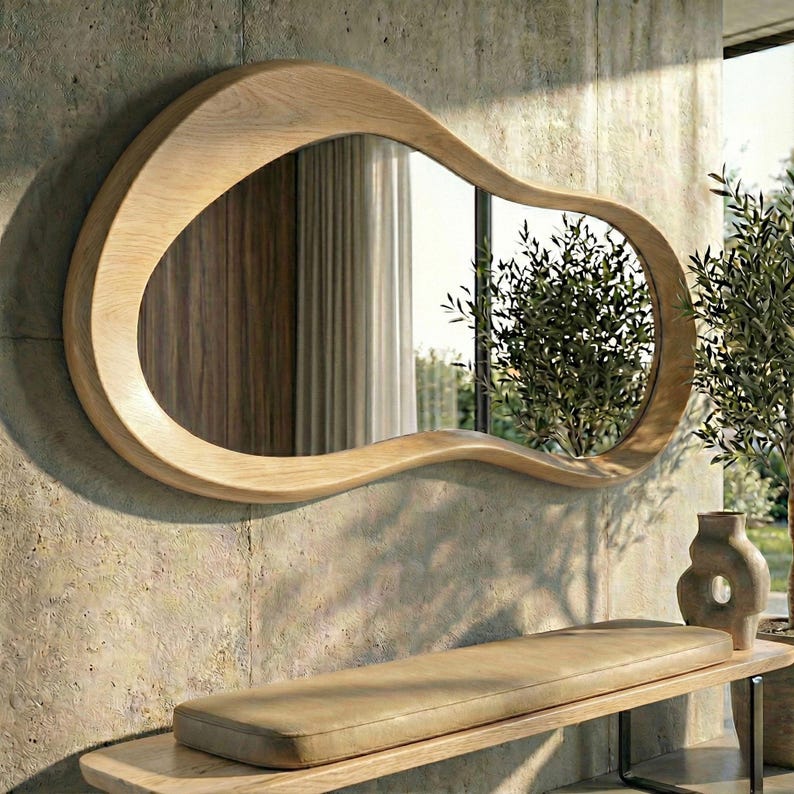 May include: A wall-mounted mirror featuring an irregular, organic, amoeba-like shape with a smooth, light-toned wooden frame. The reflective surface shows a clear view of indoor curtains and an outdoor tree, framed by the natural wood grain.