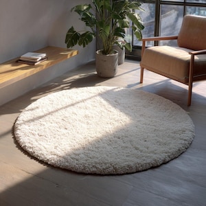 May include: A circular cream-coloured shag rug. The rug is positioned on a light wooden floor. The soft texture of the rug is visible, and it appears to be in a well-lit room, possibly near a window.