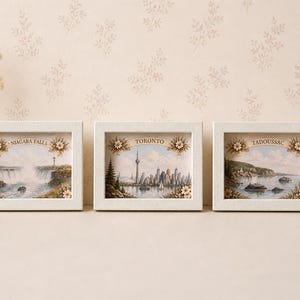 Peut inclure: Trois cadres avec des vues panoramiques de Niagara Falls, Toronto et Tadoussac. Chaque cadre blanc présente un motif floral et le nom de la ville. Les œuvres représentent des paysages avec de l'eau, des bâtiments et des éléments naturels.