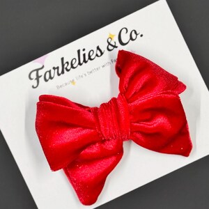 May include: A bright red velvet bow hair clip on a white card with the text "Farkelies & Co." in black script. The card is on a dark gray surface. The bow is a vibrant accessory.