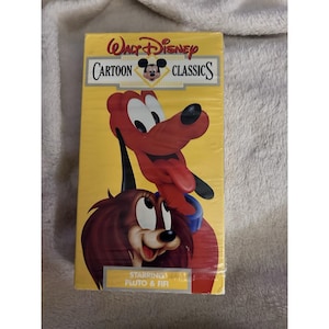 Puede incluir: Una cinta VHS de Walt Disney Cartoon Classics con Pluto y Fifi. La caja amarilla tiene el título en rojo y negro, con imágenes de dibujos animados de Pluto y Fifi. La cinta está sellada en plástico.
