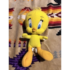 May include: A yellow plush toy of the cartoon character Tweety Bird, featuring a large round head, blue eyes with long black eyelashes, a small orange beak, and two feet. The plush has a soft texture and is depicted in a standing position.