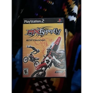 May include: A PlayStation 2 game case for MX Superfly, featuring Ricky Carmichael. The cover art depicts a motocross rider mid-air on a red and white motorcycle, with other riders and bikes in the background. The game is rated "Everyone" by ESRB.
