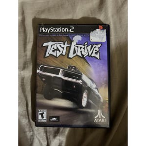 Op de afbeelding: Een PlayStation 2 game case voor "Test Drive". De cover toont een zwarte muscle car met de speltitel in een vetgedrukte lettertype. De game is beoordeeld als "Teen" en bevat het Atari-logo.
