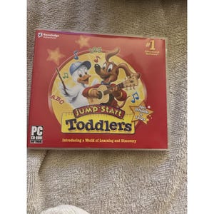 Puede incluir: Una caja de CD-ROM roja para el software educativo "Jump Start Toddlers". La portada presenta un pato y un perro de dibujos animados tocando la guitarra, con el texto "Jump Start Toddlers" e "Introducing a World of Learning and Discovery". Edades de 18 meses a 3 años.