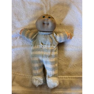 May include: A vintage Cabbage Patch Kid doll with a tan face, blue eyes, and a small smile. The doll is wearing a blue and white striped knit outfit with fuzzy white trim at the ankles. The doll's arms are outstretched.