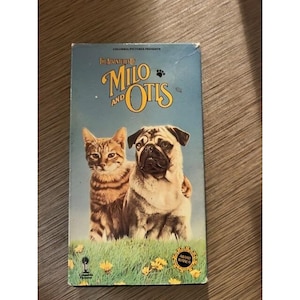 May include: A VHS tape for "The Adventures of Milo and Otis" featuring a tabby cat and a pug dog on a grassy field. The title is in yellow script with a blue background. The tape has a "Columbia Pictures Presents" logo.