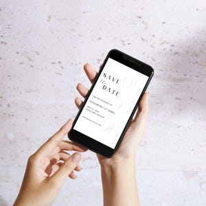 May include: A black smartphone displays a wedding "Save the Date" invitation. The text includes the date, names, and location. The phone is held in front of a textured, light-colored background.