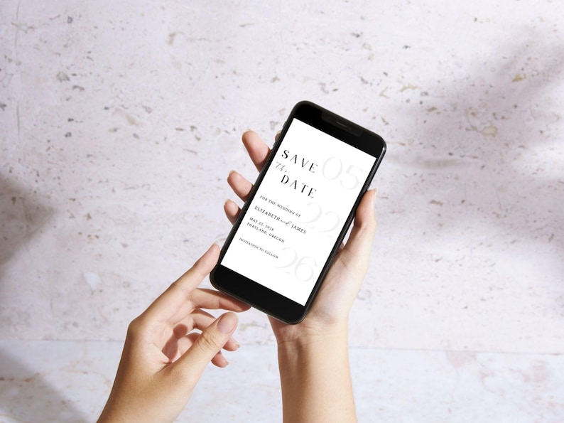 May include: A black smartphone displays a wedding "Save the Date" invitation. The text includes the date, names, and location. The phone is held in front of a textured, light-colored background.