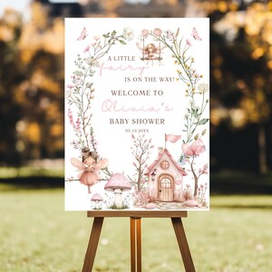 May include: A baby shower sign on a wooden easel. The sign features a fairy theme with floral accents, butterflies, and a fairy house. Text reads "A little fairy is on the way! Welcome to Olivia's Baby Shower 05.10.20XX."