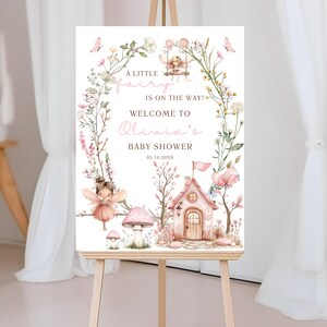 May include: A wooden easel displays a baby shower welcome sign. The sign features a watercolor illustration of a fairy theme, with a fairy house, mushrooms, and floral accents. Text reads "A little fairy is on the way! Welcome to Olivia's Baby Shower."