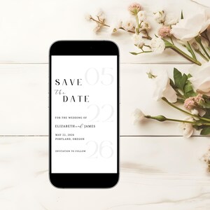 May include: A black smartphone displays a wedding "Save the Date" announcement. The text includes the date, names, and location, with floral accents. The phone rests on a white wooden surface, adorned with white and pink flowers.