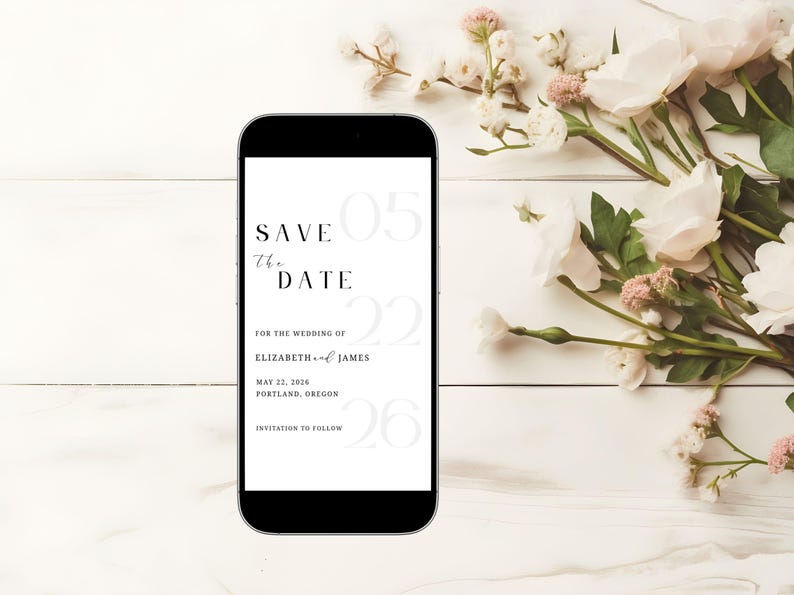 May include: A black smartphone displays a wedding "Save the Date" announcement. The text includes the date, names, and location, with floral accents. The phone rests on a white wooden surface, adorned with white and pink flowers.
