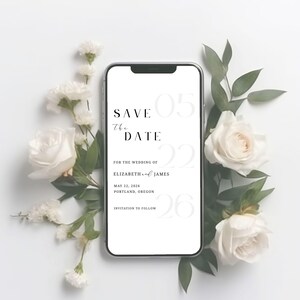 May include: A smartphone displays a wedding "Save the Date" announcement. The text includes the date, May 22, 2026, and the names Elizabeth and James, with the location Portland, Oregon. The phone is surrounded by white roses and greenery.