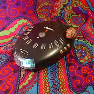 May include: A black Conair sound machine with an oval shape, featuring a central I/O button, multiple sound selection buttons labelled with text, a volume control dial, a small red indicator light, and a translucent light bar at the front.