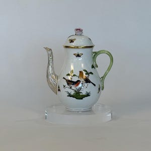 May include: White porcelain teapot with a silver spout and a green handle. The teapot features a lid with a pink rose and gold trim. The body is decorated with colorful birds, butterflies, and foliage. A decorative piece for serving tea.