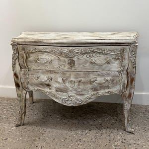 Puede incluir: Un cofre blanco envejecido de dos cajones con tallas ornamentadas y patas curvas. El cofre presenta un acabado de vetas de madera y herrajes decorativos. El estilo recuerda a los muebles antiguos franceses.