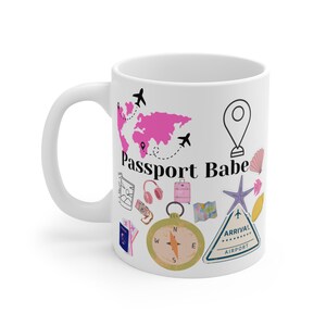 Puede incluir: Taza de cerámica blanca con ilustraciones de temática de viajes. Presenta un mapa del mundo rosa, aviones, una brújula y el texto "Passport Babe". Otros gráficos incluyen una maleta, auriculares y un mapa.