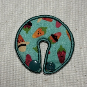 May include: A teal travel neck pillow with a repeating pattern of cartoon fruits and vegetables. The pillow has a dark green border and two teal snap closures. The pattern includes watermelon, peppers, and corn.