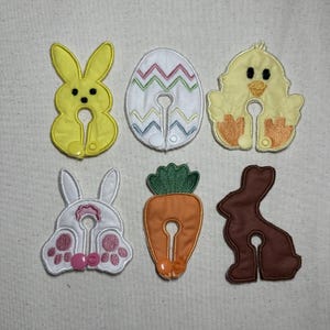 May include: A collection of six Easter-themed fabric appliques. The designs include a yellow bunny, an egg with zig-zag patterns, a yellow chick, a bunny rear, an orange carrot, and a brown bunny. Each applique has a central opening.