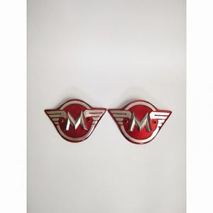 Può includere: Due emblemi metallici rossi e argentati. Ogni emblema presenta una "M" argentata stilizzata all'interno di una forma ovale rossa, con ali argentate che si estendono dai lati. Gli emblemi sono probabilmente per uso automobilistico o motociclistico.