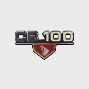 May include: A motorcycle emblem with the text "CB 100" in silver and off-white. Below the text is a red and black emblem with a silver wing design. The emblem is likely for a Honda motorcycle.
