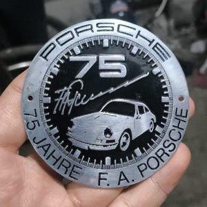 May include: A silver-colored Porsche emblem with the text "PORSCHE" around the edge. The center features the number "75", a car illustration, and the words "JAHRE F.A. PORSCHE". The emblem is held in a hand.