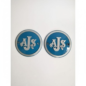 Könnte beinhalten: Zwei runde, blaue Metall-Embleme mit den Buchstaben "AJS" in silberner Schrift. Jedes Emblem hat einen silbernen Rand und zwei kleine Löcher. Wahrscheinlich für ein Fahrzeug oder eine Maschine.