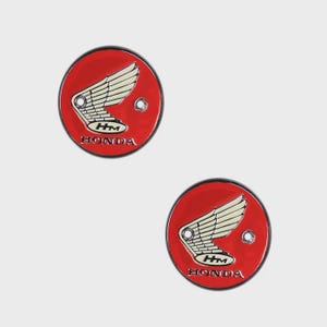May include: Two red Honda emblems with a white wing design and the Honda logo. The emblems are circular with a black border and a small hole. The background is a light gray.