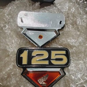 May include: Two chrome motorcycle emblems. The top emblem is a chrome plate with a diamond-shaped accent. The bottom emblem is black with the number "125" in yellow and a diamond-shaped accent with a red background and a silver wing logo.