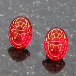 May include: A pair of red and gold earrings with a geometric design. The earrings are round and have a shiny finish.