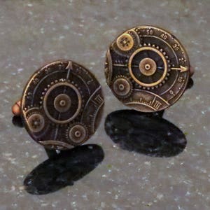 May include: Two gold-toned cufflinks with a steampunk design featuring gears and numbers. The cufflinks are round and have a detailed design.