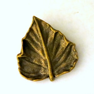 May include: A gold-toned metal leaf charm with a detailed, textured surface. The leaf is shaped like a maple leaf with a pointed tip and a central vein.