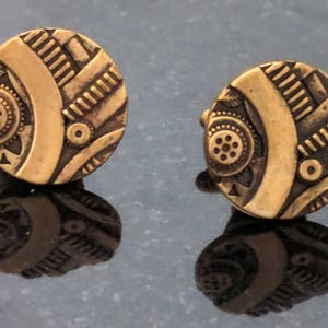 May include: A pair of round, antique gold cufflinks. Each cufflink has an intricate design with geometric patterns, including lines, circles, and a central curved element. The cufflinks are displayed on a reflective surface.
