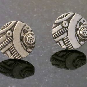 May include: A pair of silver earrings with a geometric design. The earrings are round and have a textured surface.