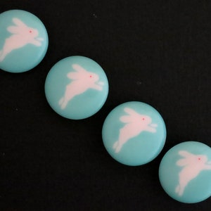 May include: Four light blue round buttons with white bunny silhouettes. Each bunny is facing right and has a pink nose.