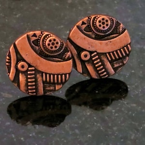 May include: A pair of copper earrings with a geometric design. The earrings are round and have a textured surface.