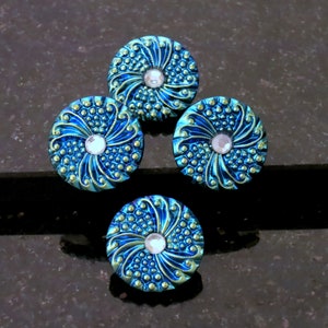 May include: Four round, ornate buttons with a teal and gold color scheme. Each button features a central, clear rhinestone and a swirling, textured design. The buttons are arranged in a square pattern on a dark surface.