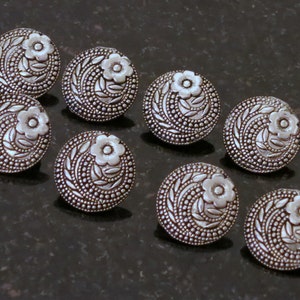 May include: Eight silver-toned buttons with a floral design. Each button has a raised flower in the center surrounded by leaves and dots.