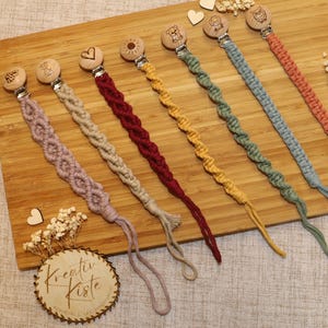 Puede incluir: Una colección de seis clips de chupete de macramé coloridos con diseños de animales y corazones de madera. Los clips están dispuestos sobre una tabla de madera, mostrando varios colores como rosa, rojo, amarillo, verde, azul y melocotón. Cada clip tiene un lazo en el extremo.